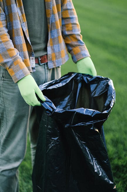 A low-angle view of a roadside rubbish pile composed of black plastic garbage bags, crumpled paper, and miscellaneous waste materials. A broken beige car tire leans against the pile on the right side,